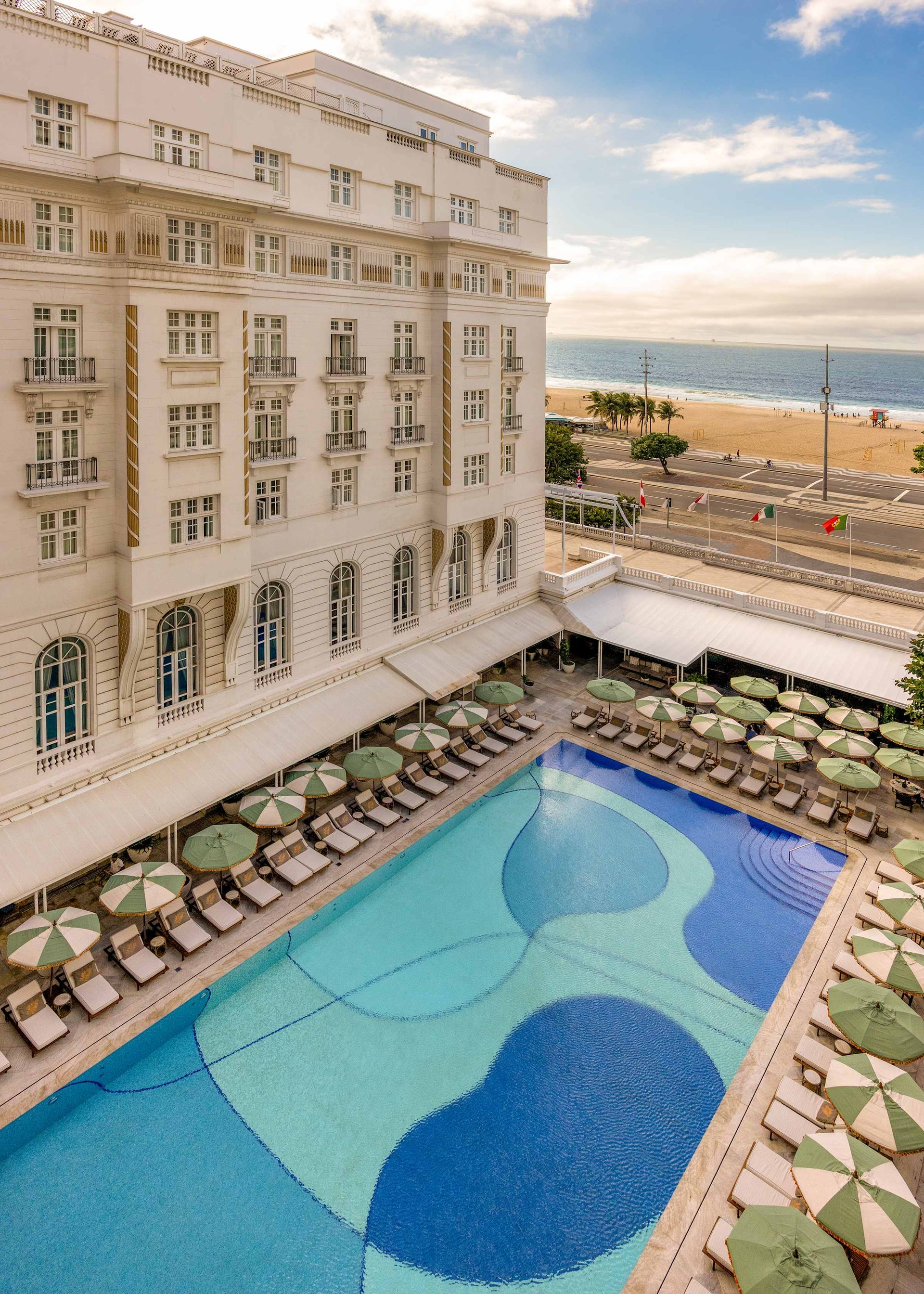 The Copacabana Palace’s pool with its blue mosaic and green parasols, the hotel’s white façade and the beach beyond